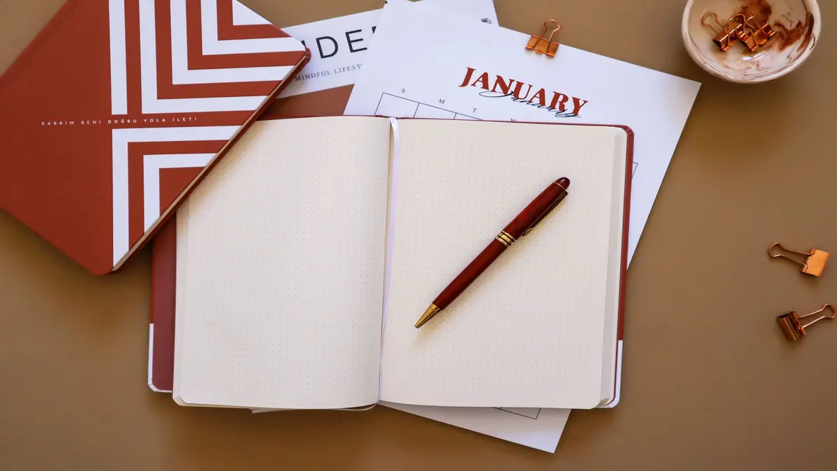 Open notebook with a pen, planner, calendar page, and office clips on a desk.