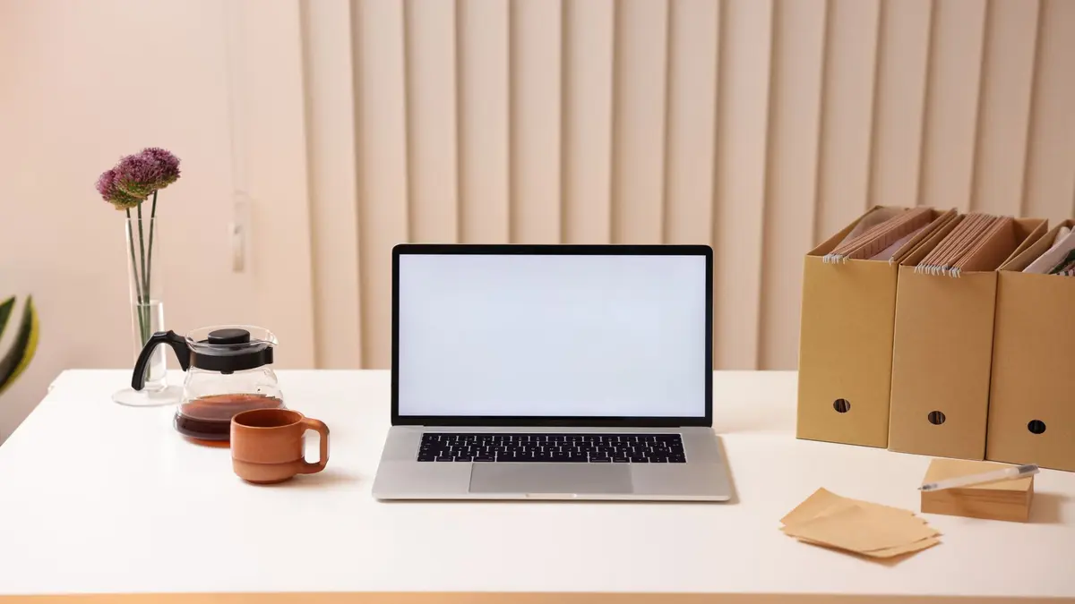 Laptop on a tidy white desk with files, coffee pot, cup, flower vase, and envelopes.